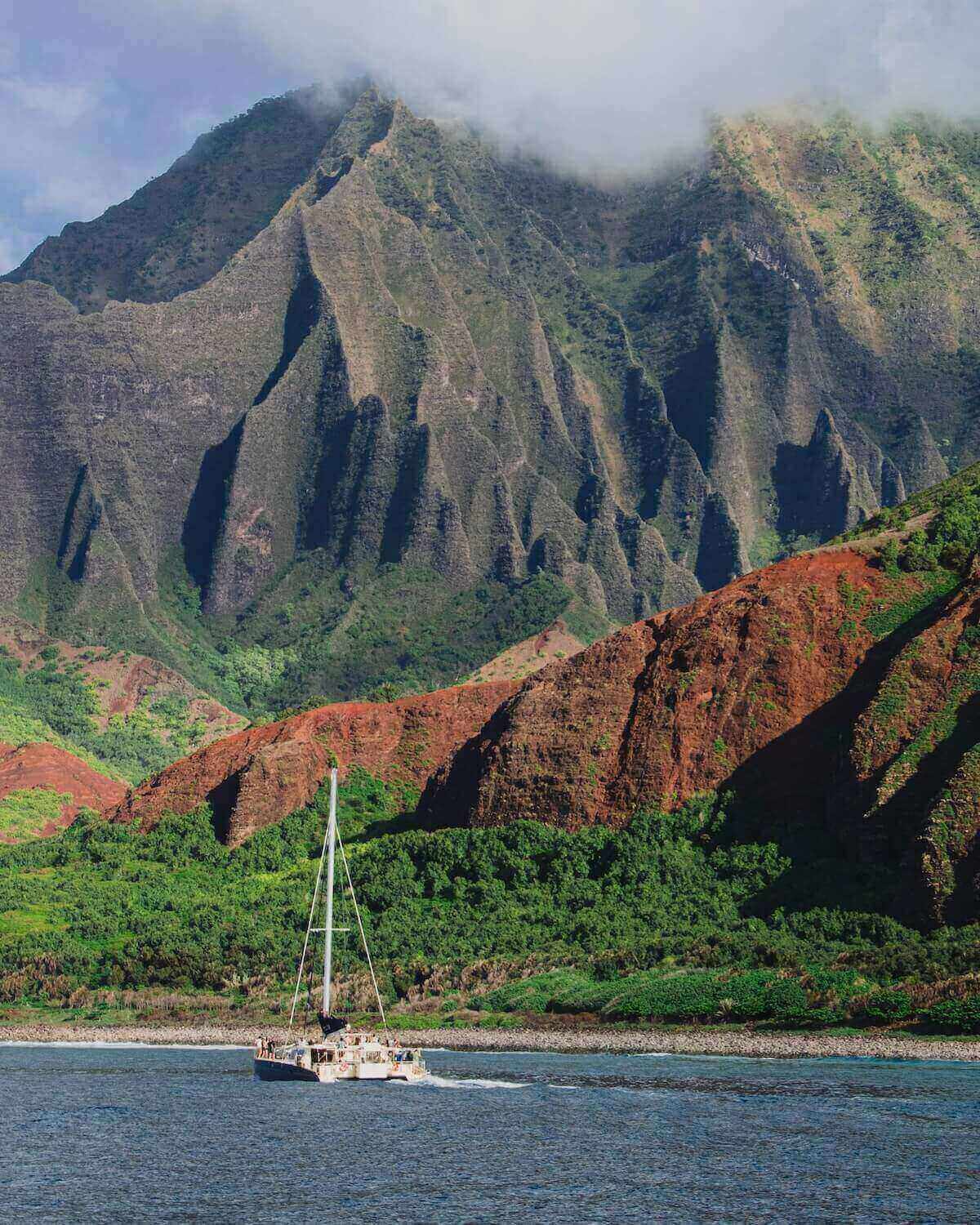 Catamaran sailing along Na Pali Coast with jagged cliffs and cloudy sky in Kauai