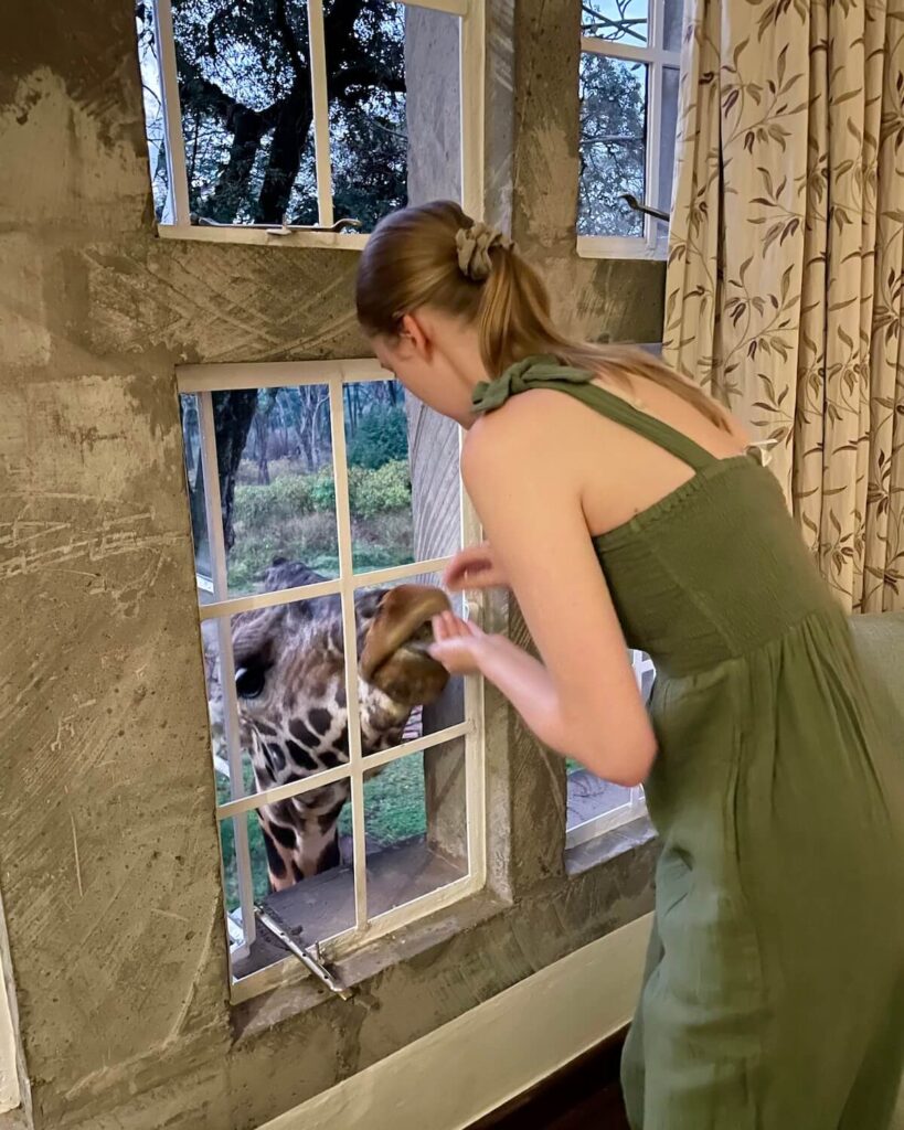 Samantha feeding giraffe through window by hand wearing green jumpsuit at Giraffe Manor