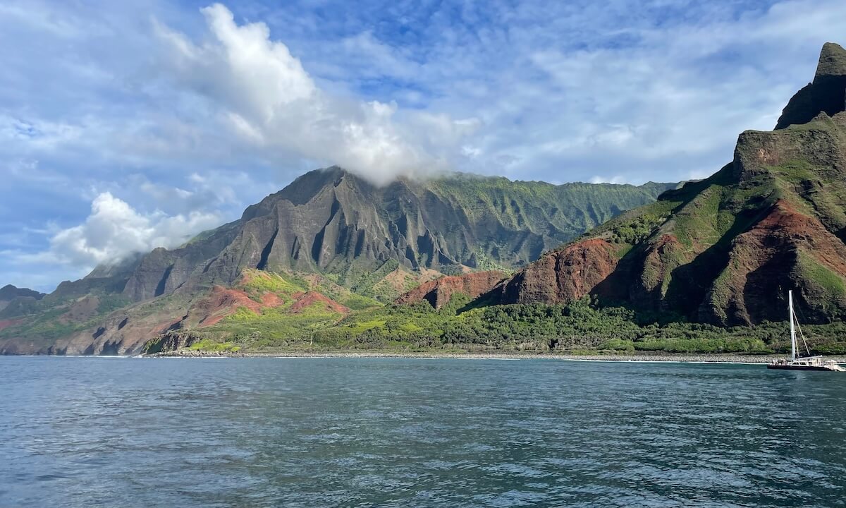 Na Pali Coast - rugged coastline covered in trees next to ocean and cloudy blue sky in Kauai