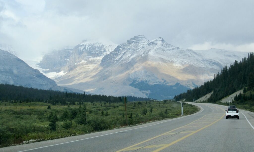 Car driving along road with impressive mountain range and grassy forest on edge in Alberta