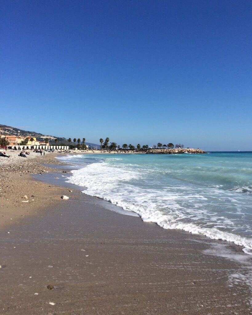 Beachfront in Menton with light blue crashing waves and bright blue sky with palm trees