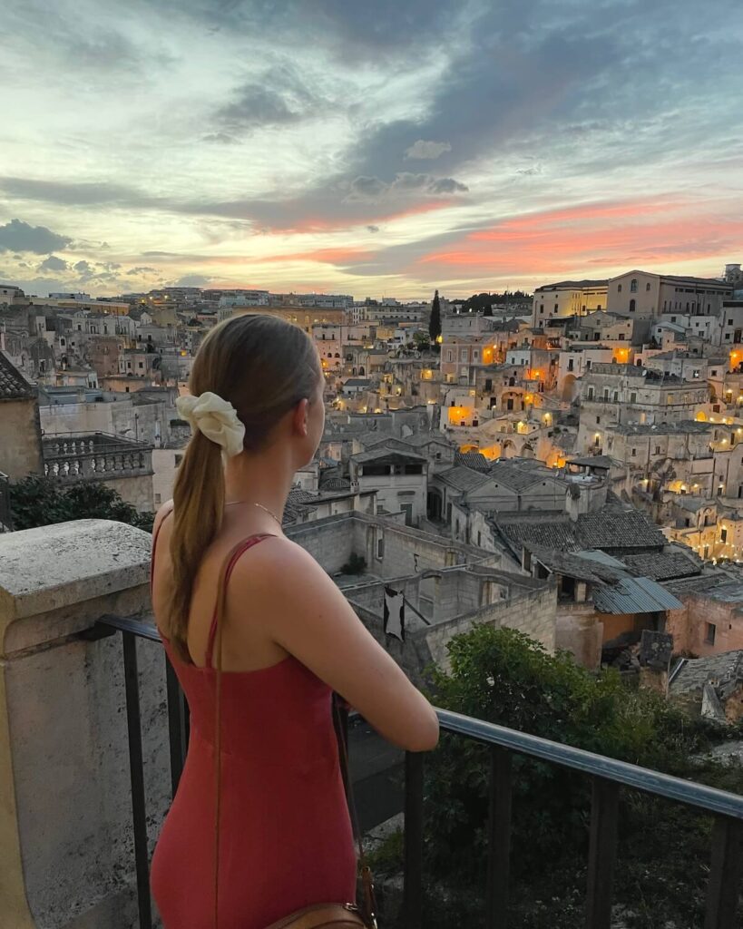 Samantha wearing pink dress looking out to Matera viewpoint at sunset with dimly lid buildings