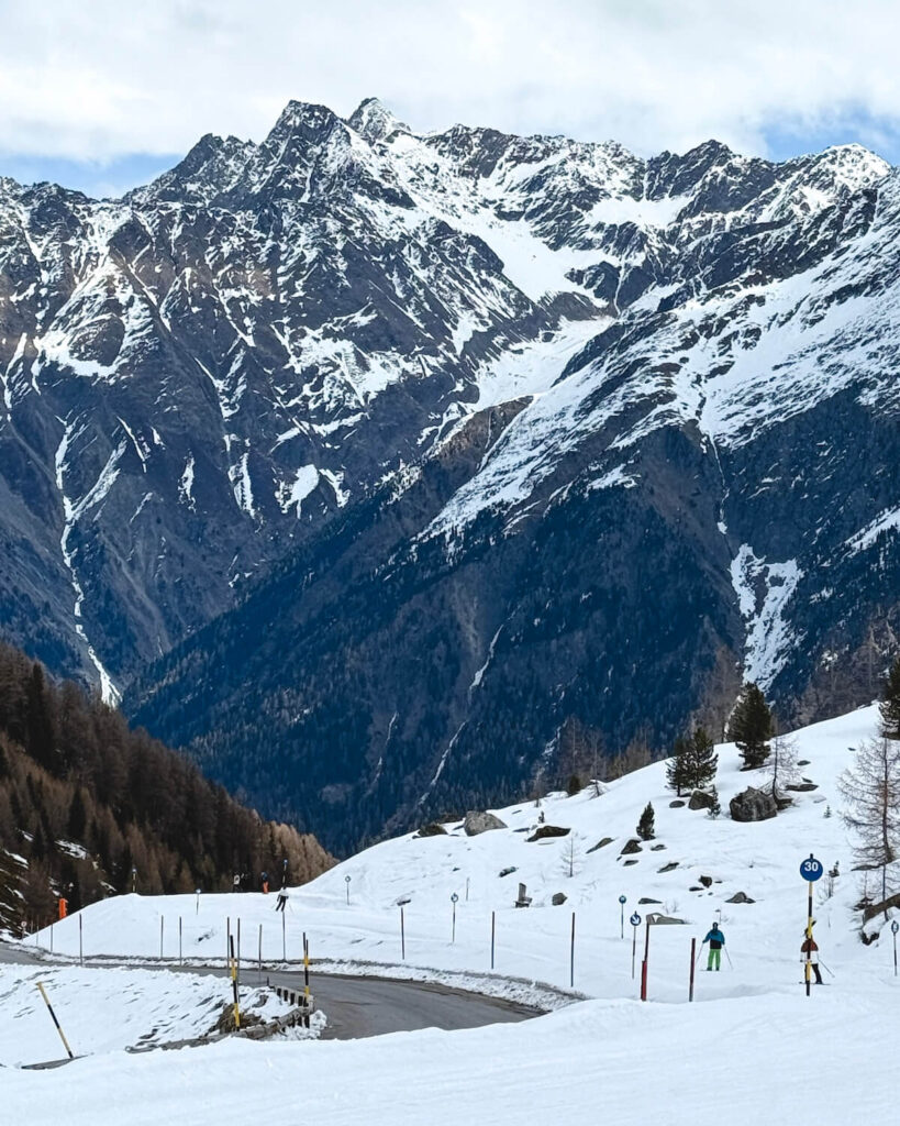 Skiers on slope next to a road with mountains ahead and green trees and rocks on side in Solden