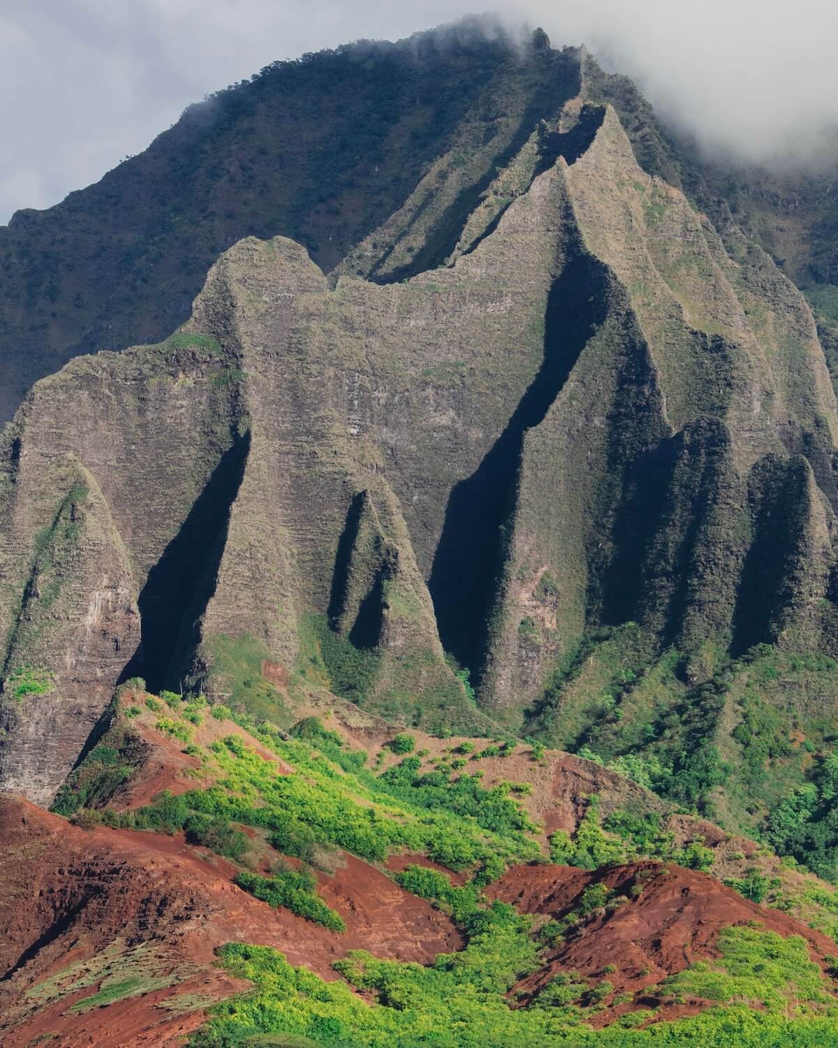 Up close view of Na Pali Coast with its sharp angles and lush green landscape below and misty clouds on top