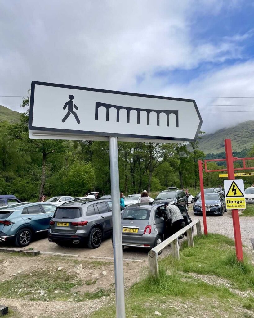 White sign with pedestrian figure walking toward a viaduct graphic - with cars parked in background
