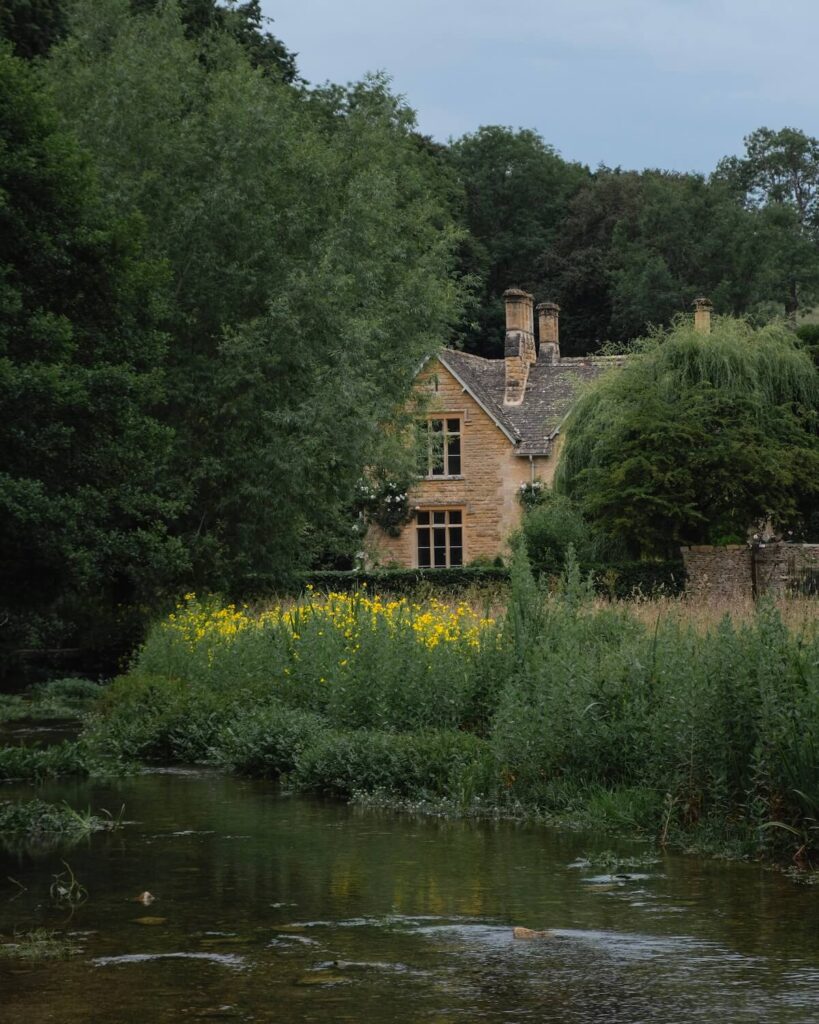 Beige brick cottage hidden behind trees and bushes and long shrubs along a stream with dim lighting
