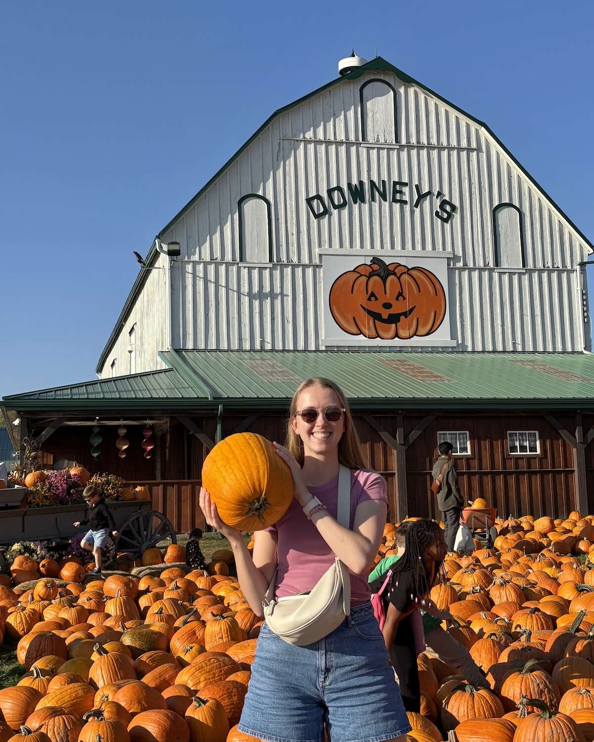Samantha holding a pumpkin surrounded by orange pumpkins with Downey's Farm house as a backdrop on sunny day