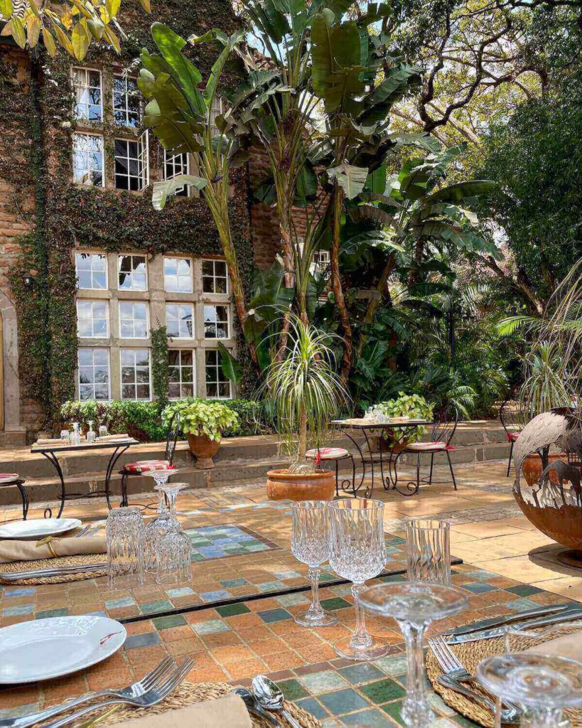 Table set for lunch with wine classes on outdoor terrace with tall trees and bushes at Giraffe Manor