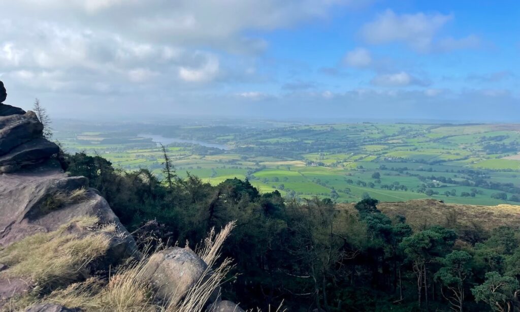 View of fields, countryside, and distant lake from The Roaches in Peak District on cloudy but bright day