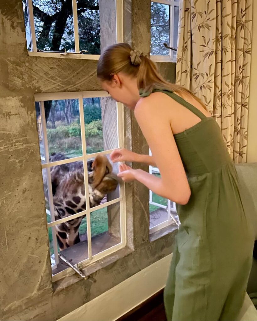 Samantha feeding a giraffe through a window in a hotel room at Giraffe Manor - wearing a green jumpsuit