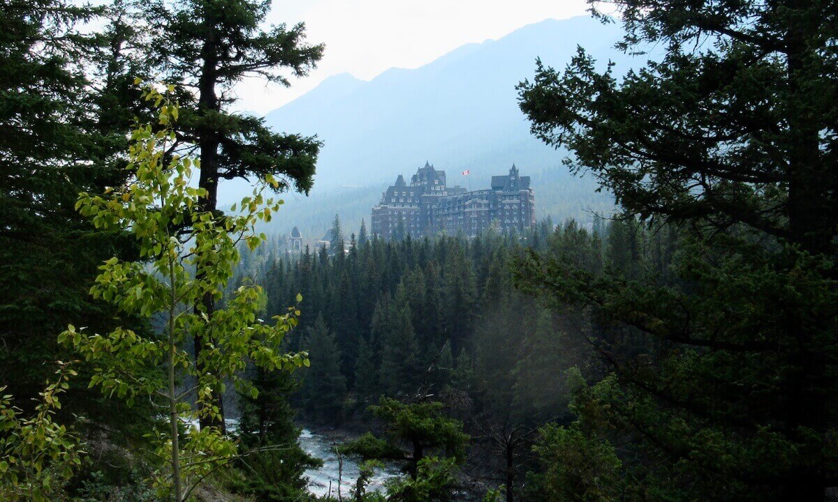 Fairmont Banff Springs Hotel perched behind pine forest and river running in front with trees framing the photo