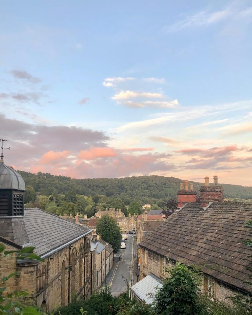 View of street in Bakewell Peak District with beige brick buildings, woods in background, and pink sunset
