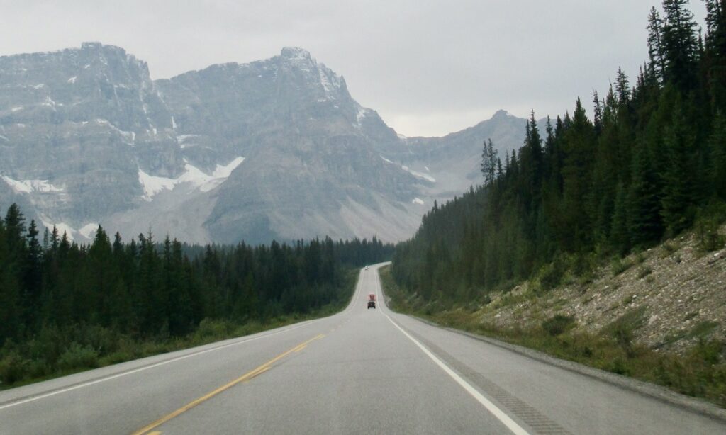 Cars driving along road with vast mountains and pine trees lining the road on a grey day in Alberta Canada