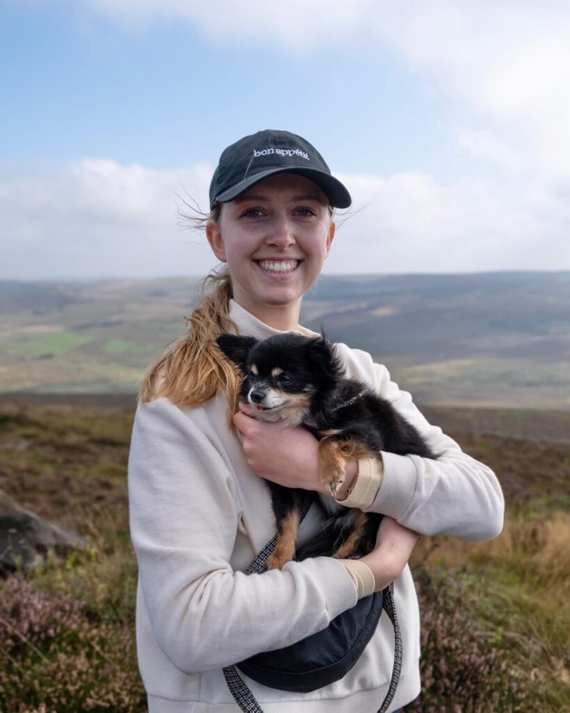 Samantha wearing baseball cap and jumper while carrying tiny chihuahua in the countryside in Peak District