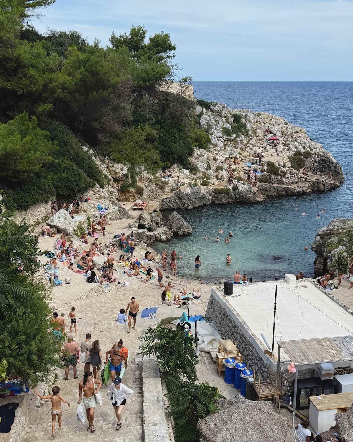 People swimming in a clear turquoise cove and sunbathing on rocky terraces beneath pine trees at Cala dell’Acquaviva