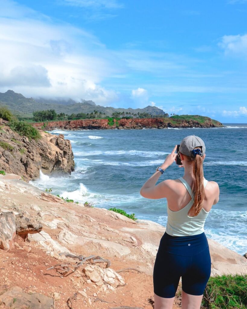 Samantha standing in active wear on Maha’ulepu Heritage Trail taking photo of the coastline in Poipu Kauai