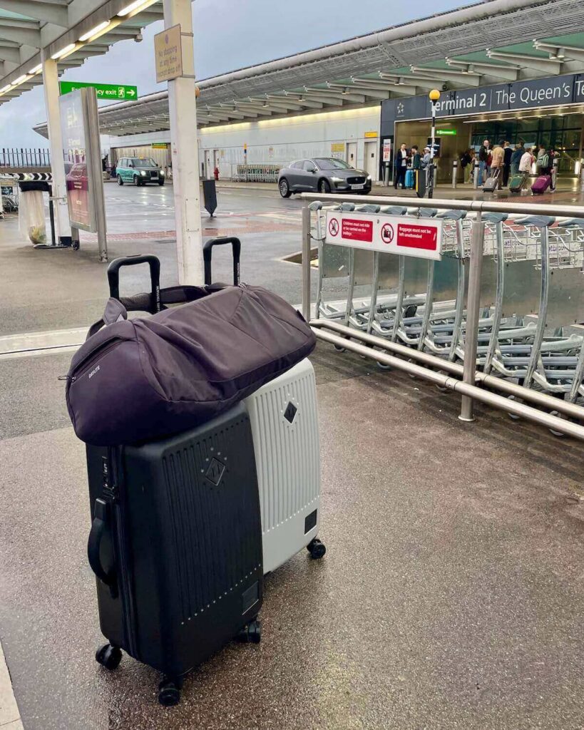 Herschel suitcase in black and grey with duffle bag on top outside Terminal 2 Heathrow Airport on wet day