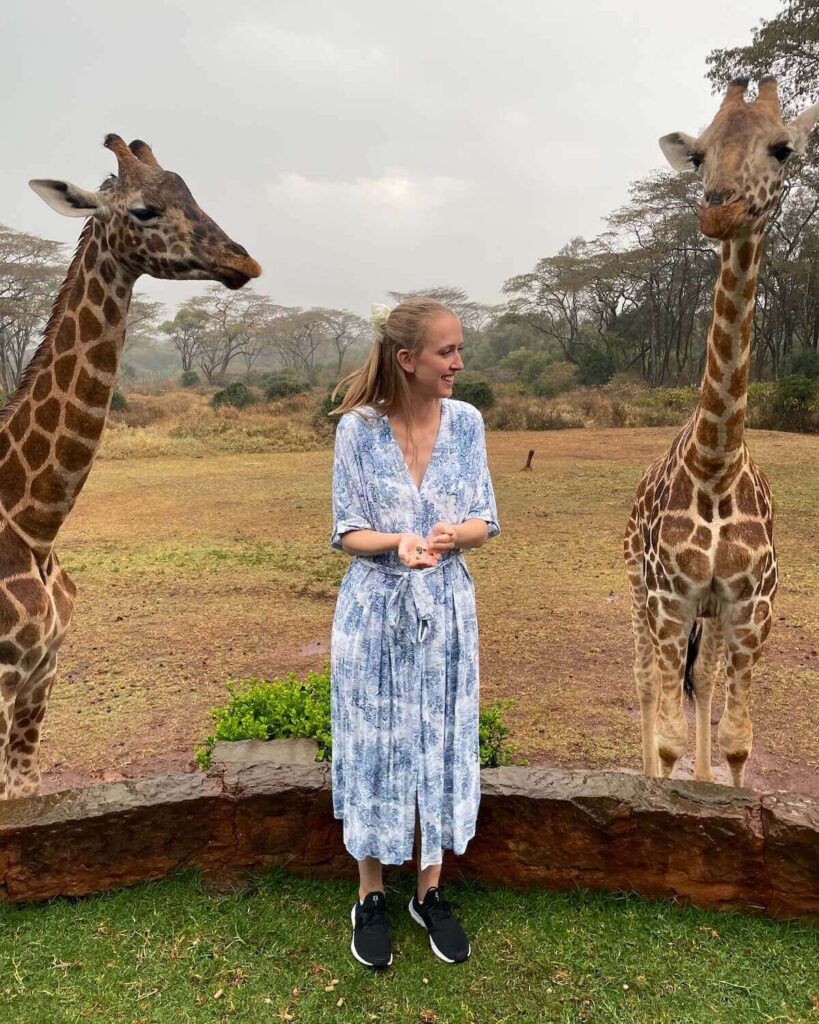 Samantha feeding 2 giraffes outside in the rain wearing a blue and white dress and black trainers on grass