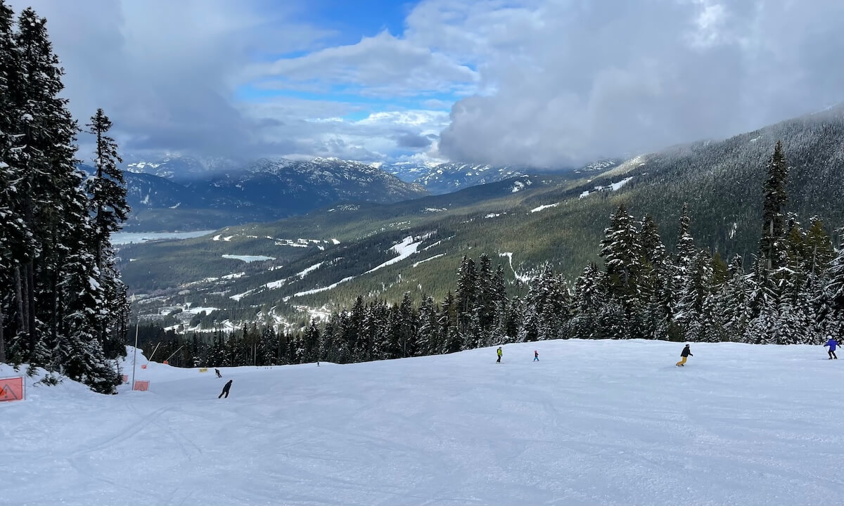 Ski run at Whistler Blackcomb with numerous snowboarders and skiers and green trees and lake in distance with clouds