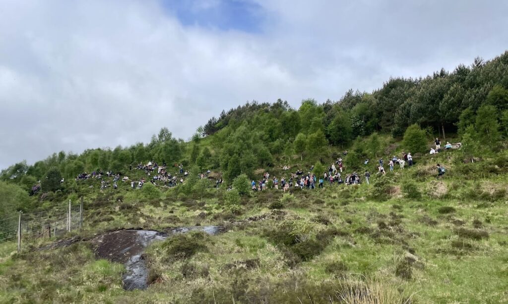 Hundreds of people sitting and standing on grassy hill to wait for the train to pass the Glenfinnan Viaduct