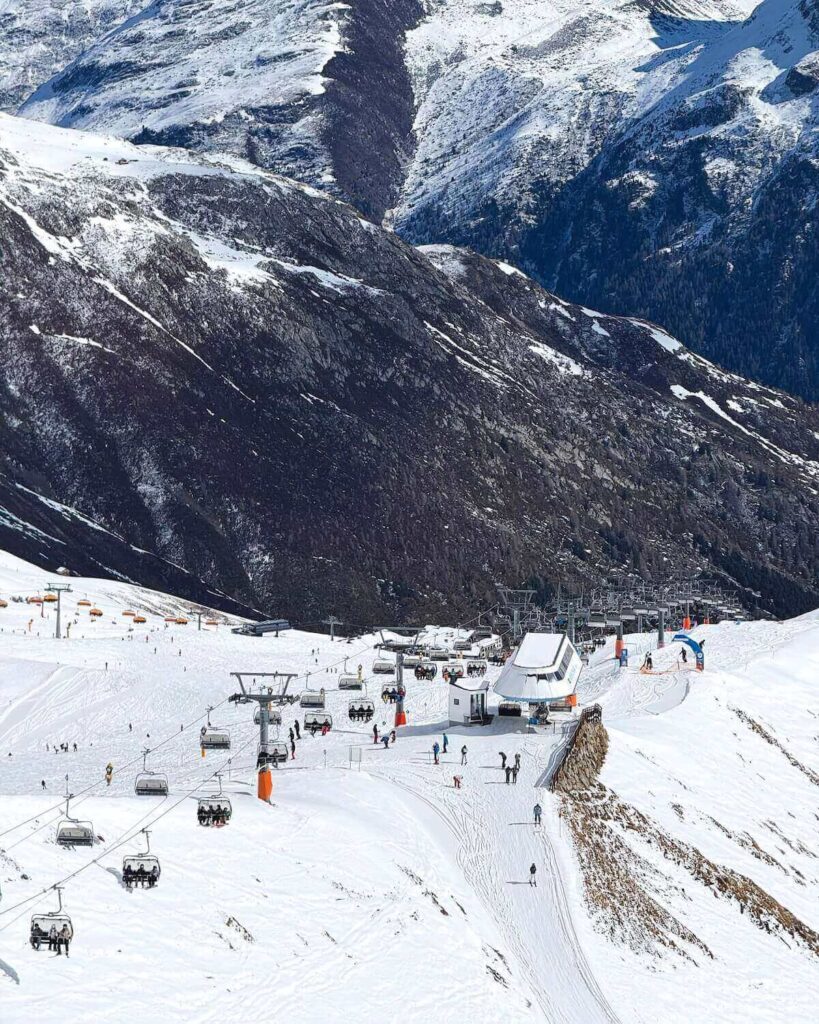 Chairlift going up ski slope in Solden with many skiers on the lift and some skiing with mountainous backdrop