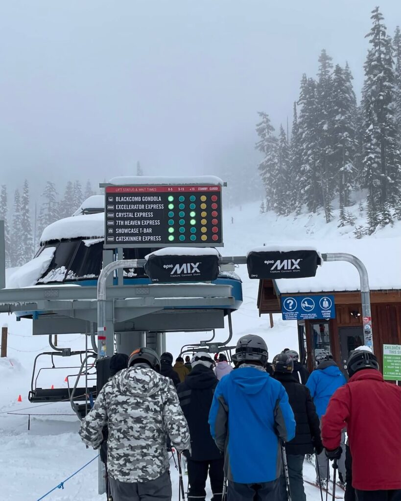 Small crowd of skiers and boarders lining up to get on lift with sign above with green lights for open lifts