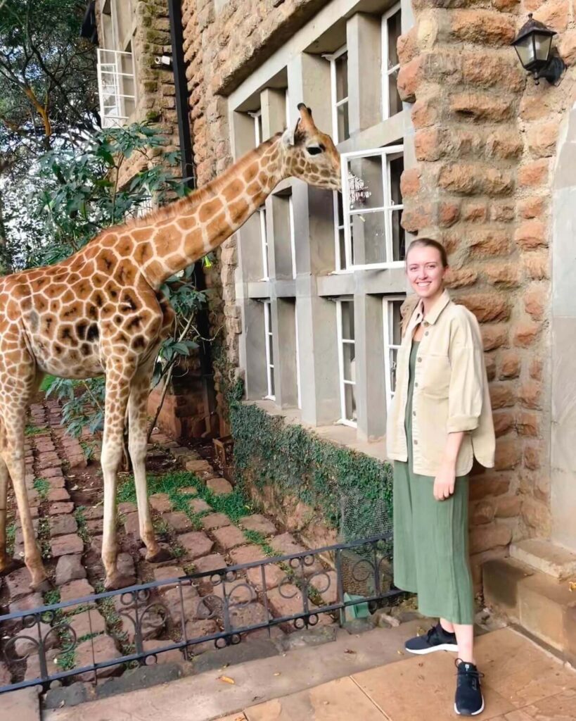 Samantha smiling while standing in front of brick building with giraffe peering into window at Giraffe Manor