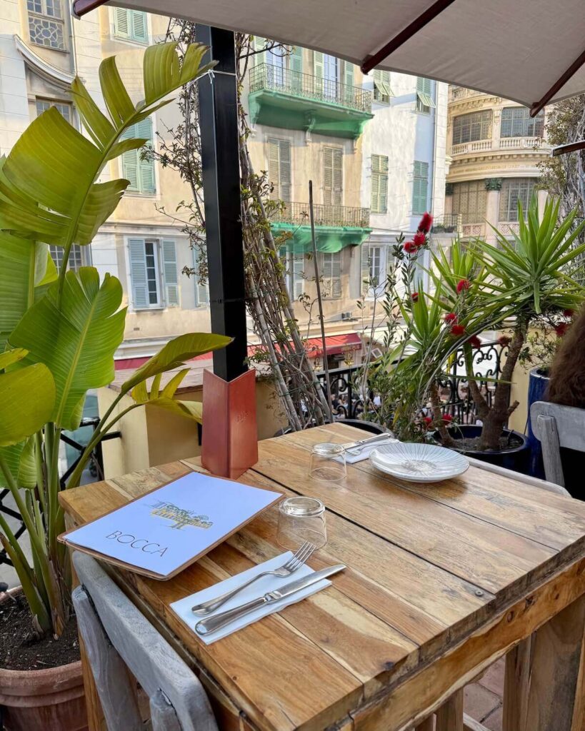 Bocca Nissa rooftop bar with exotic plants on terrace with wooden table laid for dinner and pastel buildings