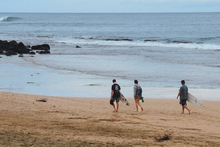 10 Days in Kauai Itinerary - 3 individuals carrying surf boards while walking along sandy beach on a grey day in Kauai
