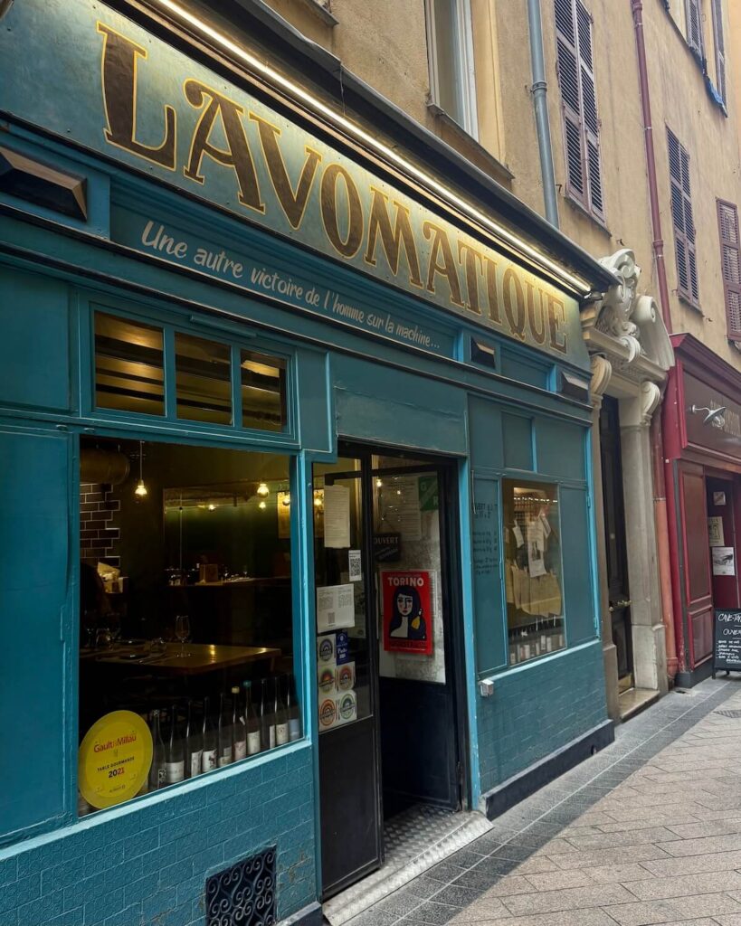 Lavomatique blue restuarant front with wine bottles in window and yellow old town buildings next door in Nice