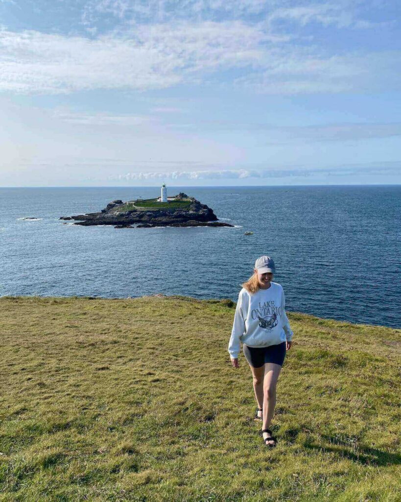 Samantha walking towards the camera on edge of cliff with ocean behind and lighthouse in the water in Godrevy Cornwall