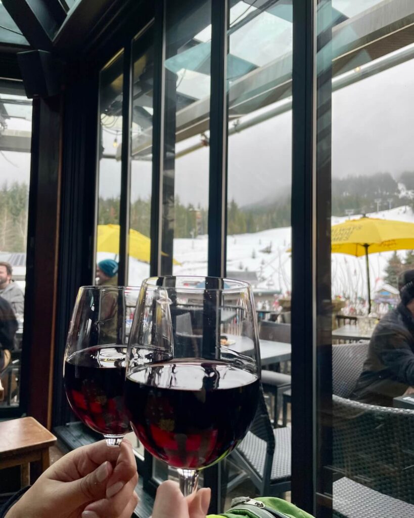 2 glasses of red wine held up against window with ski slopes and yellow umbrellas at the bottom of Whistler ski resort