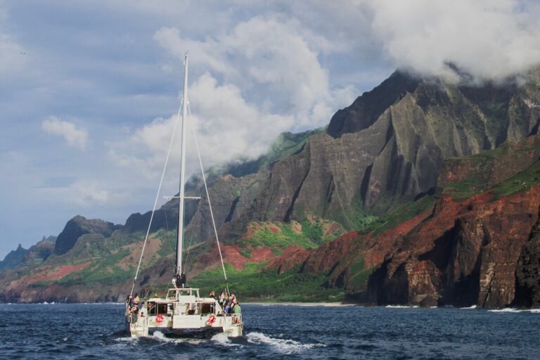 Catamaran sailing in ocean next to Na Pali coast during napali sunset cruise with vast cliffs and cloudy sky in Kauai