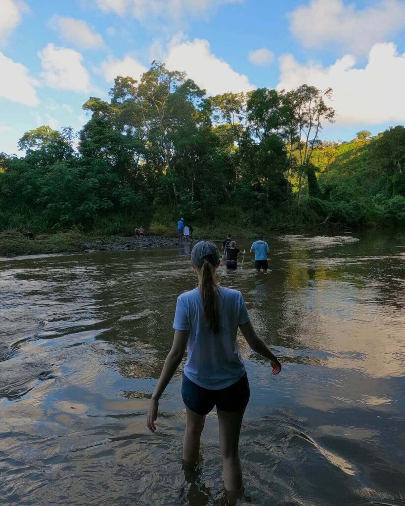 Samantha wading through stream with small group of people ahead up their thighs in water with deep forest on the other side towards Secret Falls