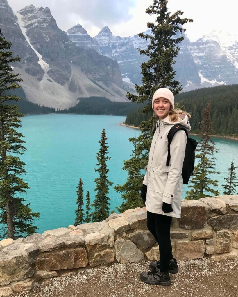 Moraine Lake - Samantha standing in front of vibrant blue green lake with skinny trees and snow-topped mountains in Banff