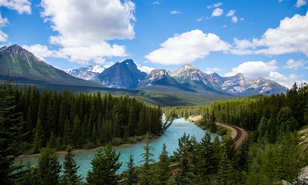 Morant's Curve - sunny view of mountains and vast green forest and a sweeping river that lines a train track in Alberta - photo by Julia Wi