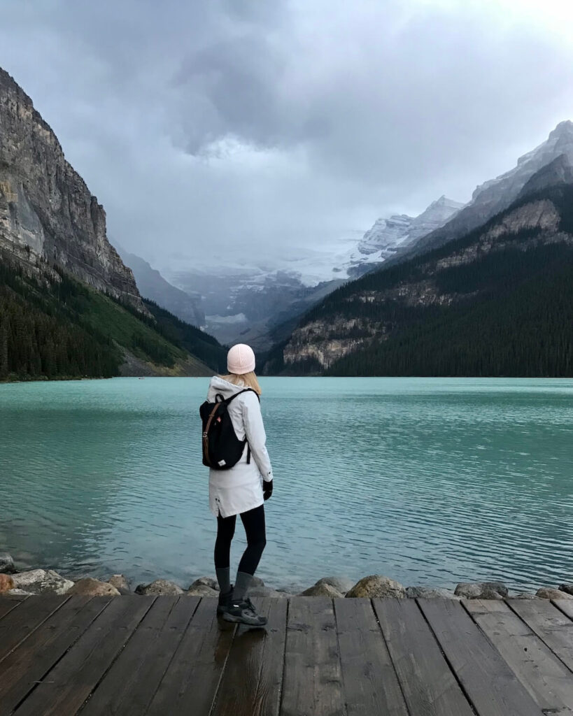 Lake Louise on a cloudy day - Samantha with her back. to the camera wearing a backpack looking at the green lake and snowy mountains in Banff