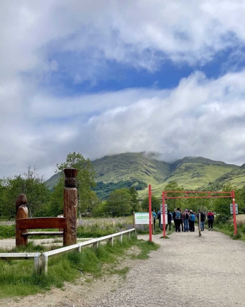 Group of walkers standing near to red arch way on path to Glenfinnan Viaduct Trail with green mountains in background