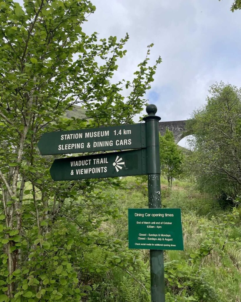 Green signs pointing in the direction of Viaduct Trail and Viewpoints and Station Museum, sleeping & dining cars with glimps of viadcut in distance