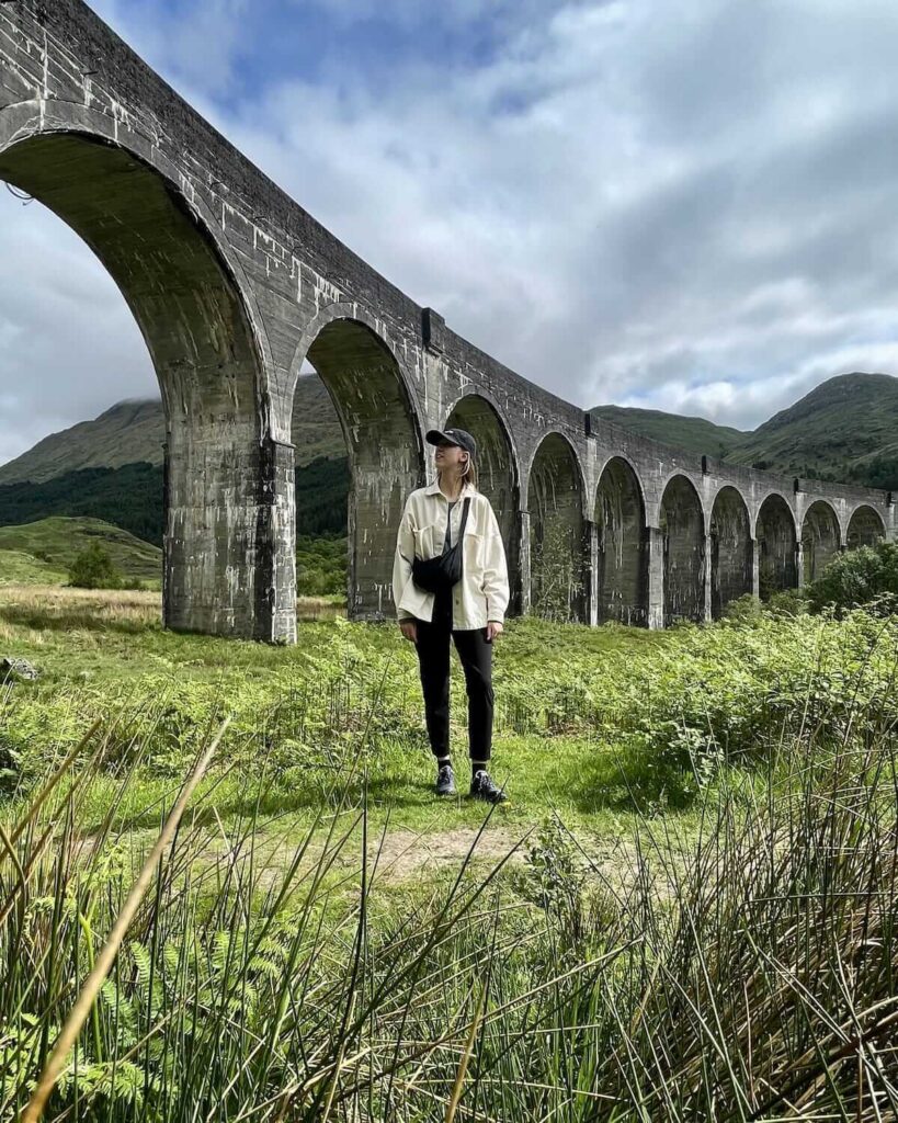 Samantha wearing cream jacket and black trousers standing in field in front of Glenfinnan Viaduct with grasses at forefront of camera