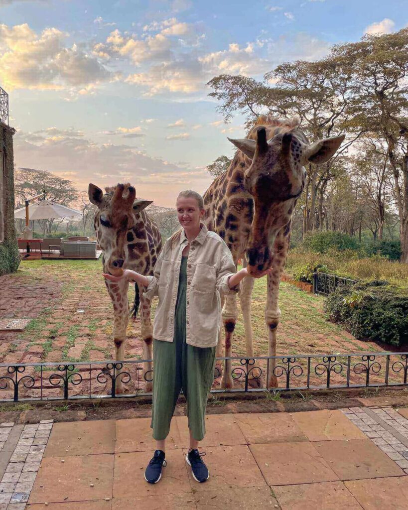Giraffe Manor - me standing next to 2 giraffes and feeding them from my hands wearing a green jumpsuit and sunrise clouds