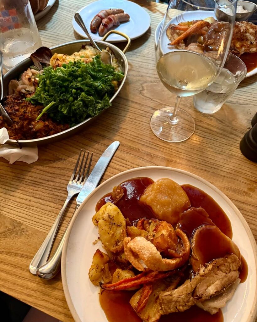 Roast dinner covered in gravy with wine glass, plate of sausages and waters on wooden table from the Chequers pub