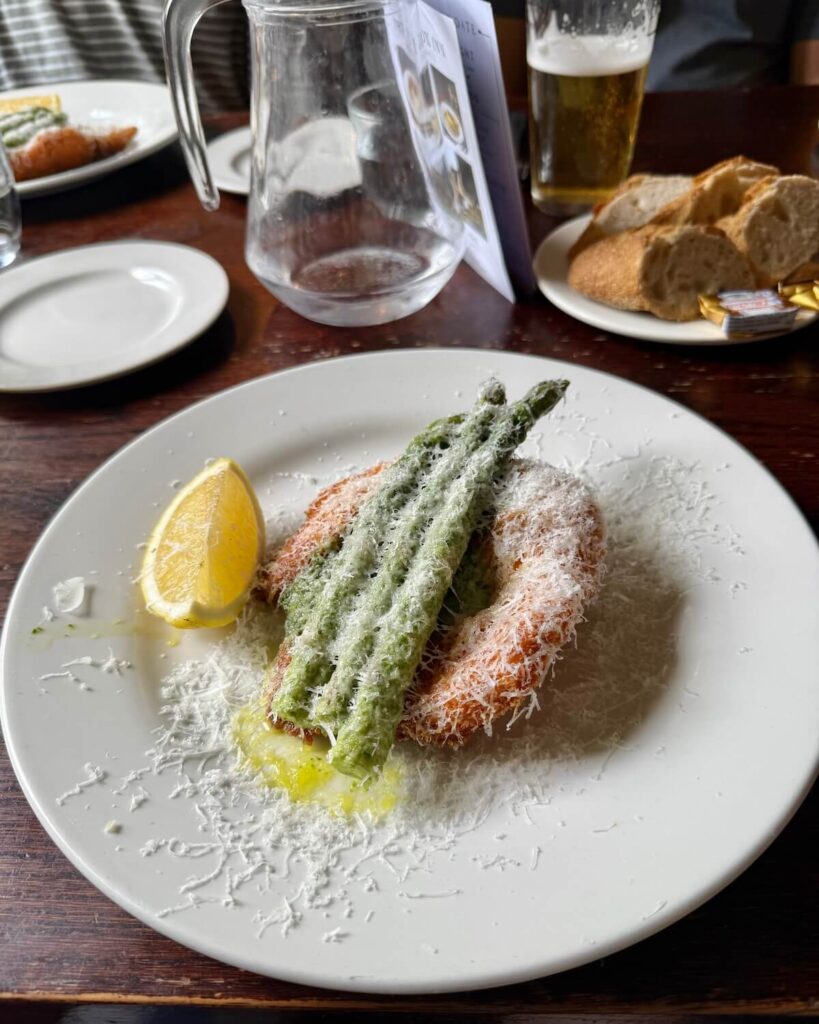 Breaded chicken with asparagus and parmesan topped on a plate with bread and beer on table at The Fox Inn Broadwell