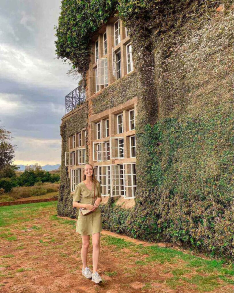 Samantha wearing short green dress and grey trainers and smiling while standing in front of vine covered Giraffe Manor building
