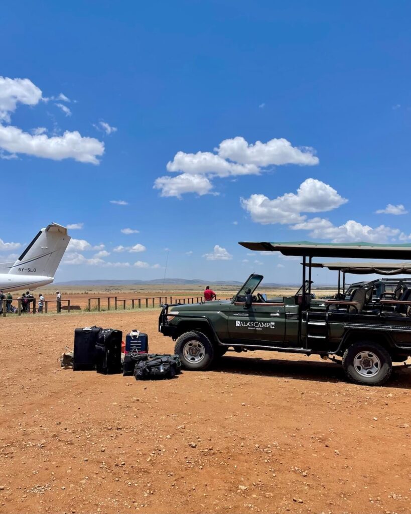 Safari vehicle on dirt path in front of Masai Mara landing strip with blue sky and small aircraft tail in background and safari luggage on the ground