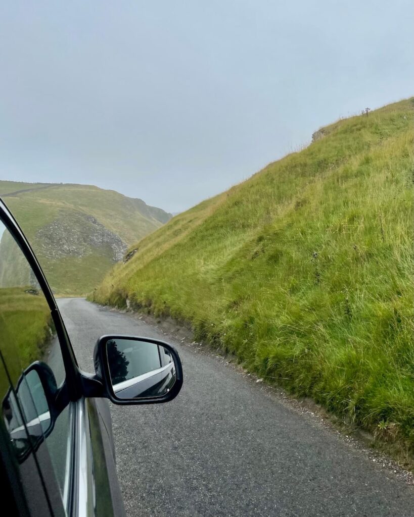 Taken out of car window while driving Winnats Pass with grey sky and grassy hills alongside road in the Peak District