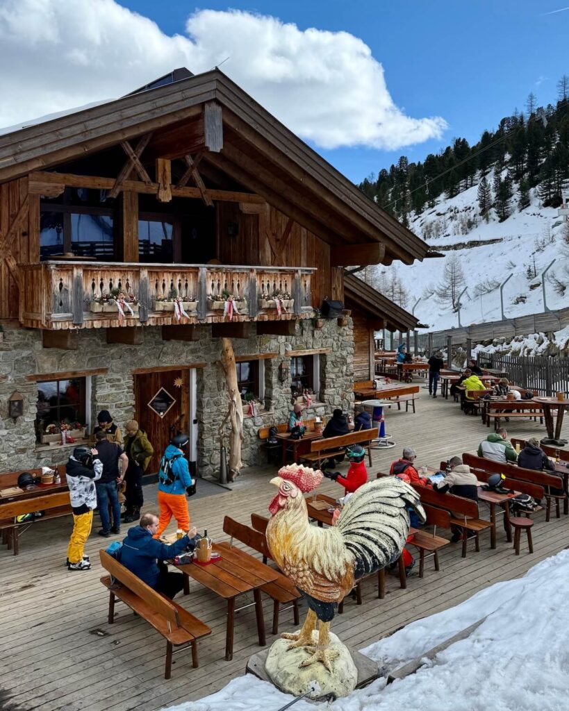 Annemaries Hühnersteign exterior of restaurant with rooster statue in forefront and busy terrace with people on picnic ables on sunny day in Solden
