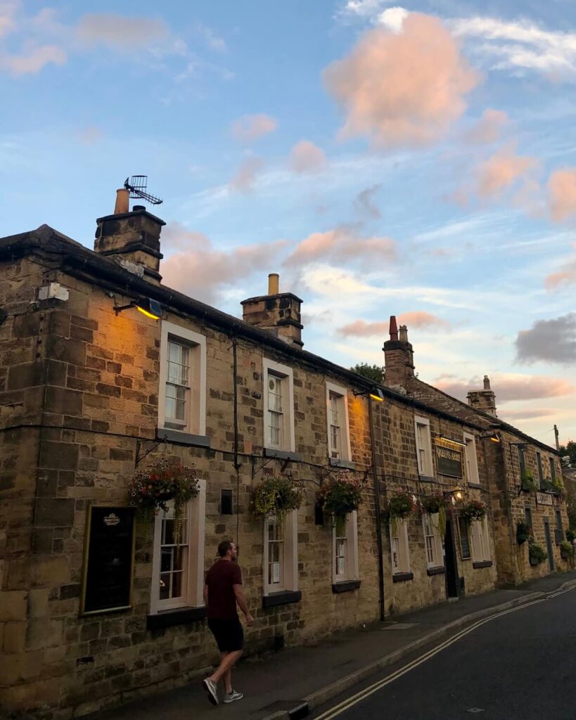 Row of quaint brick houses on high street with hanging baskets of flowers and golden hour lighting with man walking down street in Bakewell Peak District