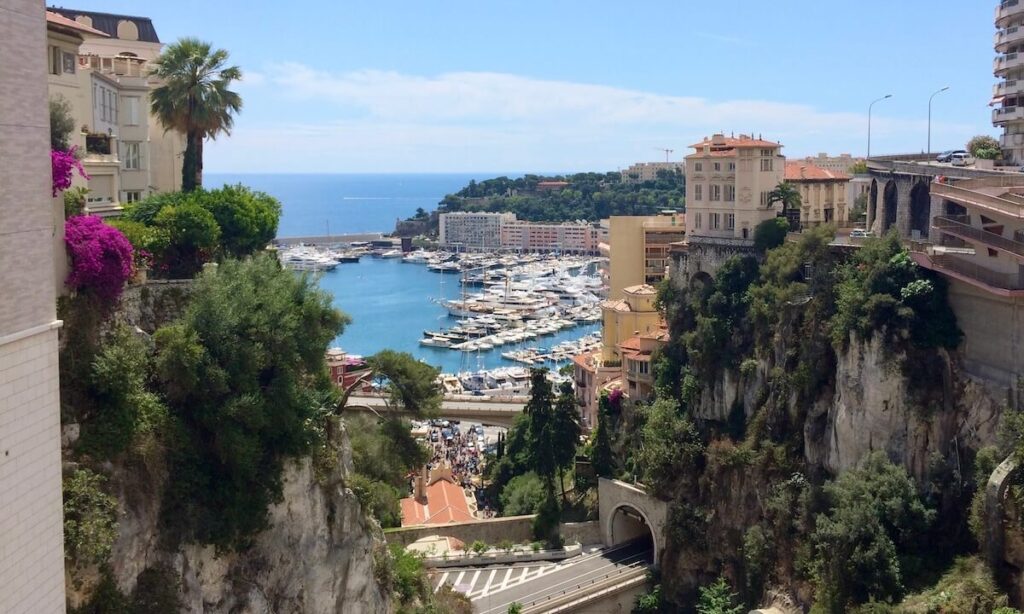 Monaco port view from train station with yachts on blue water, flowers and plants growing from rocky walls and yellow buildings on cliff edges