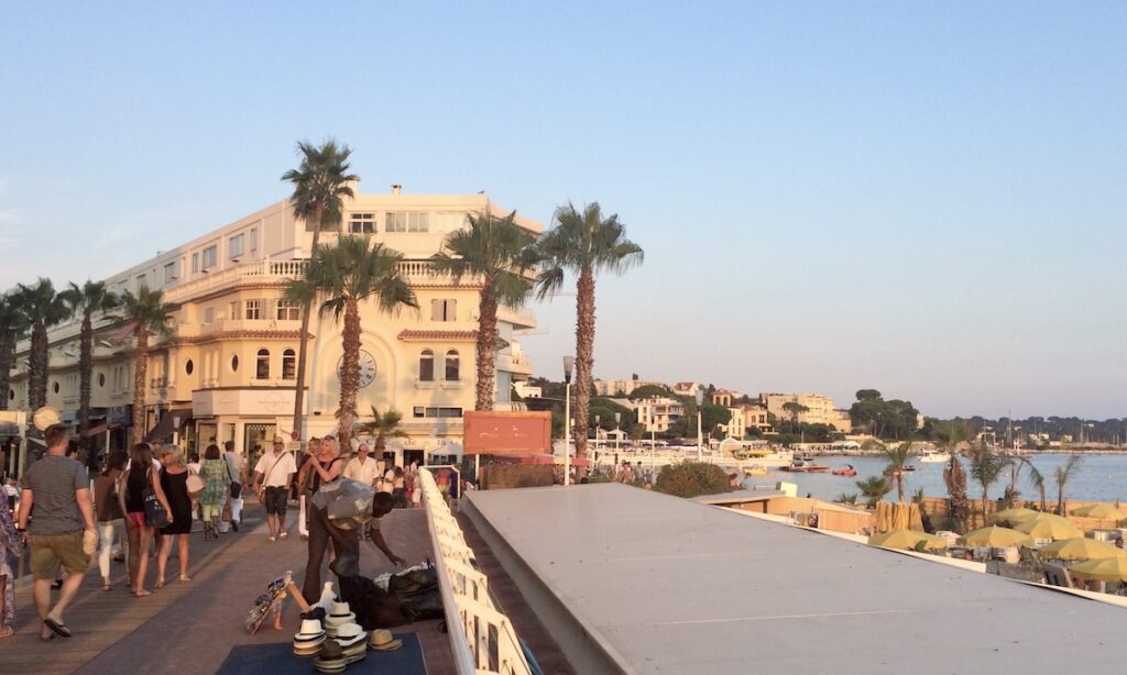 Boulevard Édouard Baudoin in Juan-les-Pins at golden hour - yellow building with large clock - flanked by palm trees and busy with pedestrians